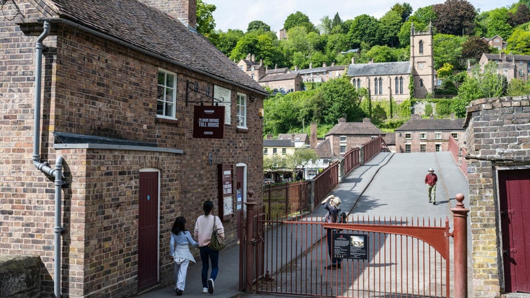 A view with a redbrick Toll house to the left and metal brown gates that seal the entrance across the famous Ironbridge Gorge. A couple of people are stopping to walk through the gates and others are looking at the Toll House. In the distance are green leafy trees and an old church.